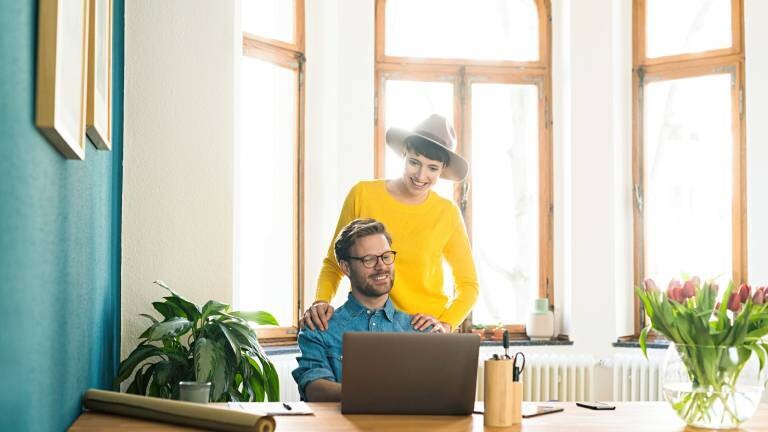 Ein entspanntes Unternehmerpaar arbeitet im Homeoffice am Laptop und lächelt. Das Arbeitszimmer ist mit Blumen geschmückt.