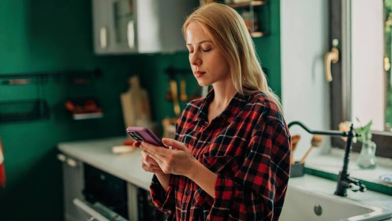 Eine Frau lehnt sich an ihre Küchenablage und vertreibt sich die Zeit mit einem Blick aufs Smartphone