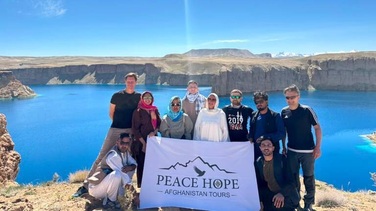 ARD-Korrespondenten Markus Spieker (hintere Reihe erster links) macht für eine Doku mit einer Reisegruppe Urlaub in Afghanistan. Die Gruppe posiert für ein Foto auf einem Fels, im Hintergrund Wasser und gebirgige Landschaft. Sie halten einen weißes Transparent vor sich mit der Aufschrift: Peace Hope - Afghanistan Tours.