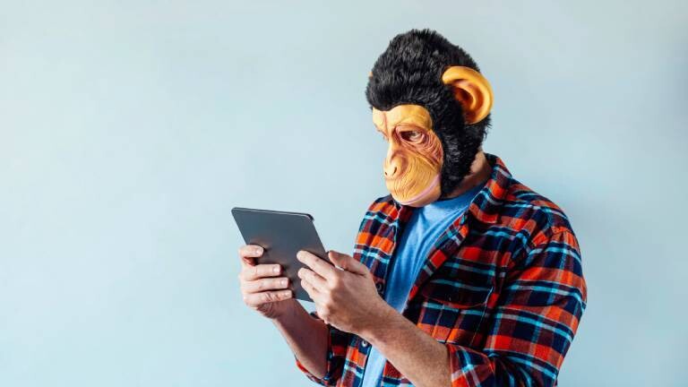 Ein Mann trägt eine Affenmaske und starrt auf ein Tablet, das er in der Hand hält. Er steht vor einer hellblau gestrichenen Wand.