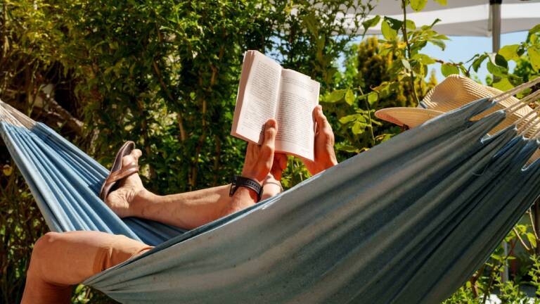 Eine Person mit Sonnenhut liegt in einer Hängematte und liest ein Buch.