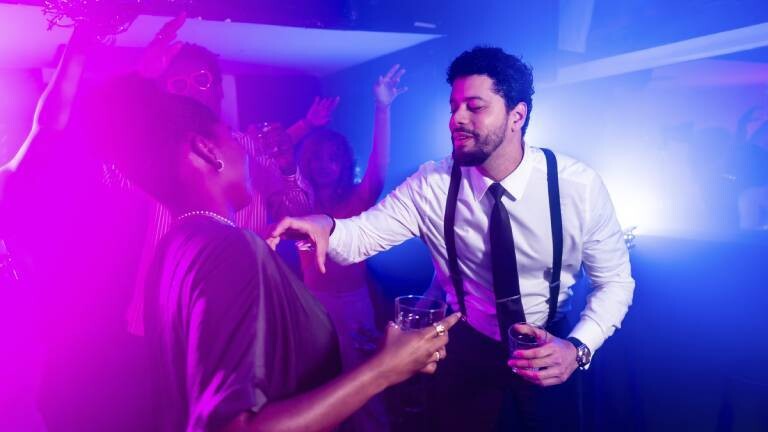 Mann flirtet mit Frau, während er alkoholische Getränke in der Hand hält und auf einer Nachtclubparty mit Freunden im Hintergrund tanzt