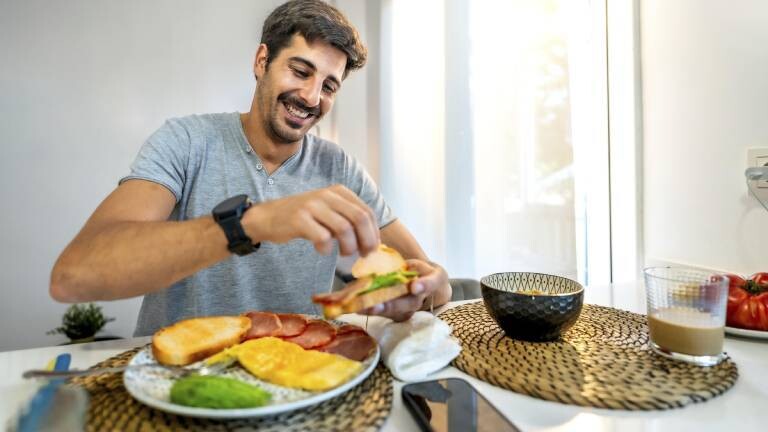 Ein Mann sitzt in der Küche am Frühstückstisch. Vor ihm stehen verschiedene Lebensmittel wie Obst, Brot, Müsli. Zu trinken hat er einen Milchkaffee. Der Mann belegt sich gerade ein Brot. Sein Handy liegt neben dem Teller.