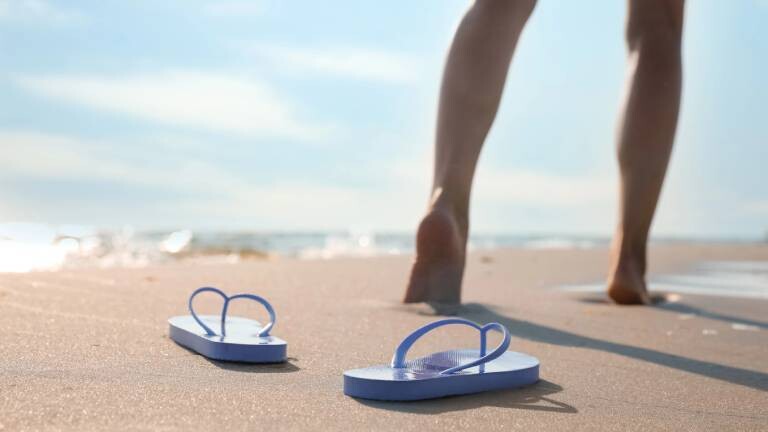 Frau läuft barfuß am Strand, links der Ozean, vorn im Bild ihre Flip-Flops