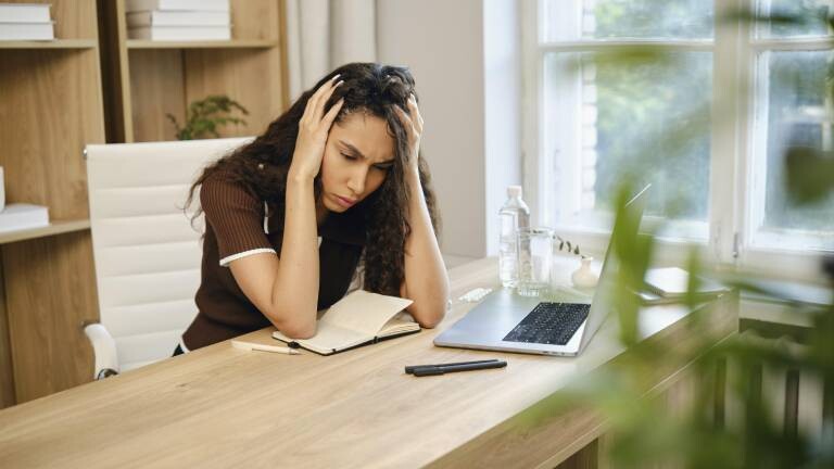 Eine müde junge Frau sitzt an ihrem Schreibtisch, sichtlich gestresst und stützt den Kopf in die Hände. Der Arbeitsplatz ist mit einem Notebook, einem Laptop und einer Wasserflasche ausgestattet