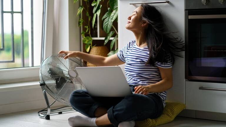 Asiatische Frau sitzt mit Laptop auf gekreuzten Knien auf dem Boden und genießt den Wind eines elektrischen Ventilators.