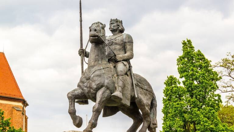 Bronzenes Reiterstandbild von Bolesław I. (dem Tapferen) vor einem bewölkten Himmel, flankiert von grünen Bäumen und dem Dach eines Backsteingebäudes.