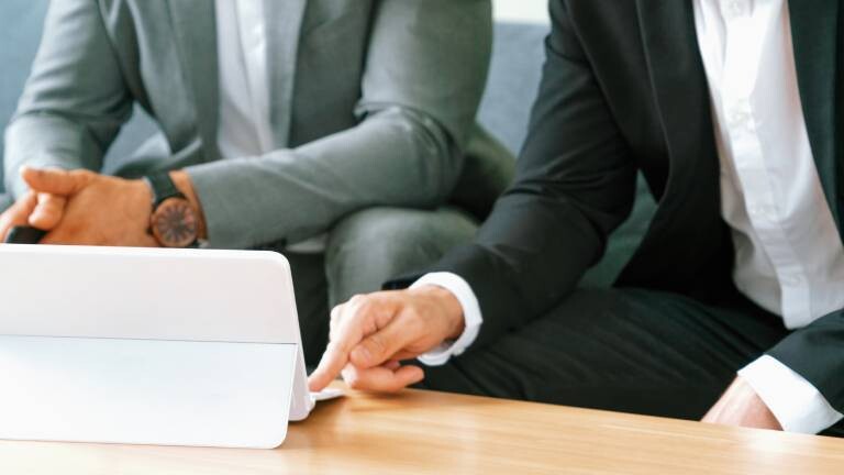 Zwei Personen in Business-Kleidung sitzen nebeneinander an einem Holztisch. Ihre Gesichter sind abgeschnitten. Eine Person im schwarzen Anzug deutet mit dem Zeigefinger auf den Bildschirm eines weißen Tablets, das vor ihnen steht. Die andere Person im grauen Sakko schaut zu und hat die Hände im Schoß verschränkt.