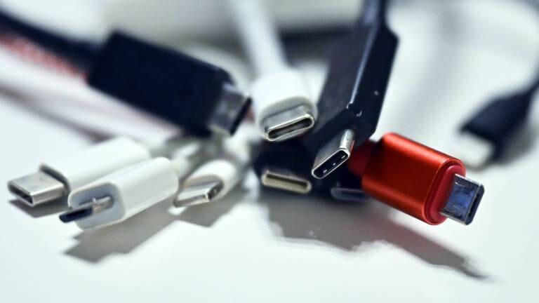 Ein Bündel verschiedener Ladekabel liegt ungeordnet auf einer weißen Oberfläche. Im unscharfen Vorder- und Hintergrund sind Stecker wie USB-C, Micro-USB und Lightning zu sehen. Ein roter Micro-USB-Stecker auf der rechten Seite sticht farblich hervor und ist scharf fokussiert.