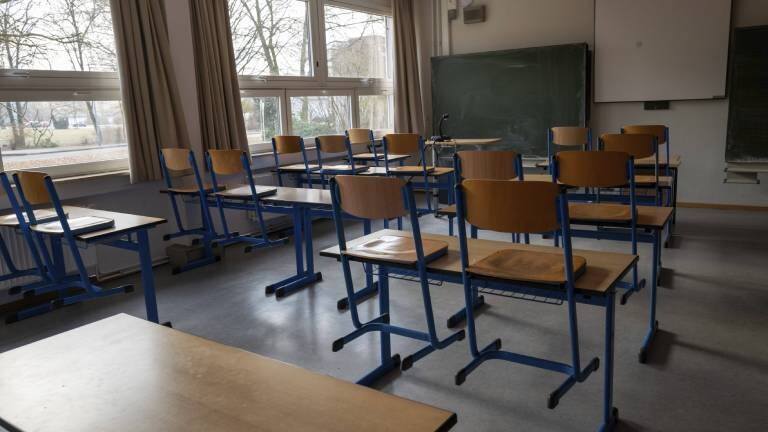 Ein leerer, tageslichtdurchfluteter Klassenraum mit hölzernen Schultischen und Stühlen mit blauen Metallbeinen. Im Hintergrund befinden sich eine Kreidetafel und eine weiße Projektionsfläche an der Wand.