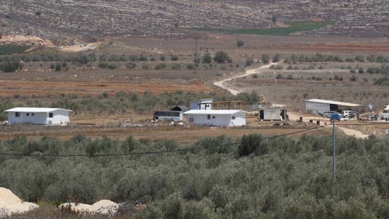 Ein kleiner, neu errichteter israelischer Siedlungsaußenposten mit weißen Fertighäusern und Schuppen im Westjordanland, umgeben von Olivenbäumen vor kargen Hügeln.