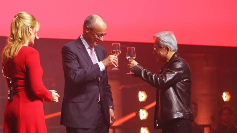 Bundeskanzler Friedrich Merz im dunklen Anzug und Nvidia-Chef Jensen Huang in seiner typischen schwarzen Lederjacke stoßen auf einer Bühne mit Weingläsern an. Links steht eine Moderatorin in einem roten Kleid mit dem Rücken zur Kamera, der Hintergrund ist in kräftigem Rot beleuchtet.