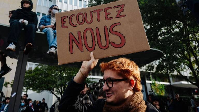 Ein junger Mann mit Brille und Schal hält bei einer Demonstration in Paris ein Pappschild mit der Aufschrift „ECOUTEZ NOUS“ (Hört uns zu) in die Höhe. Im Hintergrund sitzen weitere Protestierende auf einem Unterstand, während im unteren Bildbereich eine Menschenmenge vor einem Gebäude zu sehen ist.