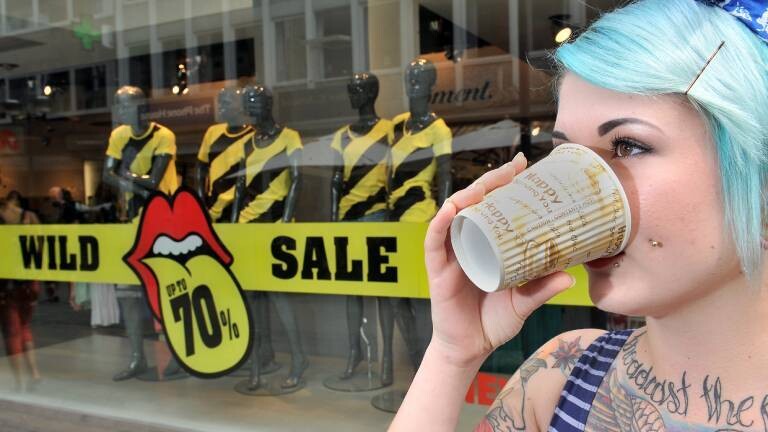 Eine Frau steht in der Fußgängerzone vor einem Schaufenster, trinkt Kaffee aus einem Pappbecher, auf dem happy shining you gedruckt ist. Im Schaufenster steht Wild Sale up to 70 %.