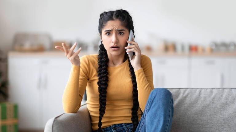 Eine Frau sitzt auf dem Sofa im Wohnzimmer und telefoniert mit einem Scam-Anrufer.