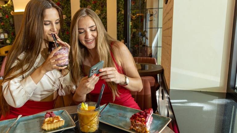 Zwei Frauen sitzen im Cafè, trinken Drinks. Auf dem Tisch steht das bestellte Gebäck. Sie freuen sich, weil sie was lustiges im Smartphone sehen.