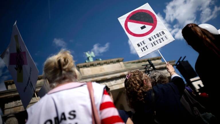 Demonstration gegen Rechtsextremismus vor dem Brandenburger Tor in Berlin; im Fokus hält eine Person ein Schild mit der Aufschrift „NIE WIEDER IST JETZT!“ unter einem durchgestrichenen Hitler-Schnitt.