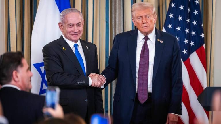 Benjamin Netanjahu und Donald Trump reichen sich die Hand, nachdem der israelische Premier ankündigte, dem Friedensplan zuzustimmen. Im Hintergrund die Flaggen Israels und der USA. Reporter fotografieren die Szene.