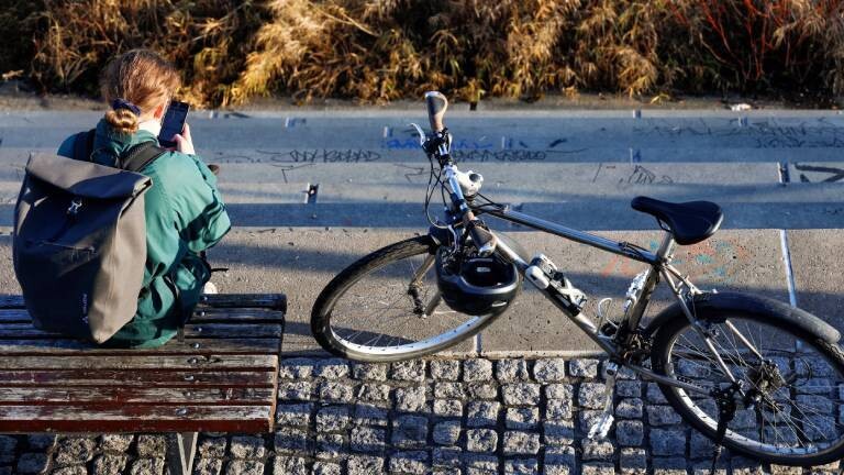 Eine Frau sitzt am Elbe-Ufer auf einer Bank. Sie trägt eine Jacke und Rucksack. In der Hand hält sie ihr Smartphone. Neben ihr steht ihr Fahrrad.