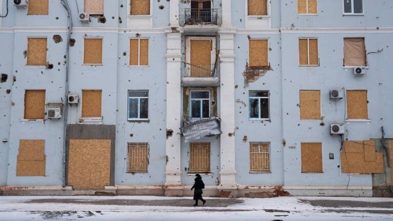 Kramatorsk, Oblast Donezk, Ukraine: Ein Wohnhaus in Kramatorsk, dessen Fenster vernagelt und dessen Fassade von Granatsplittern und Kleinwaffenfeuer gezeichnet ist, steht beschädigt in einer ostukrainischen Stadt, während ein Zivilist im Winter inmitten der andauernden Invasion Russlands in der Ukraine vorbeigeht.