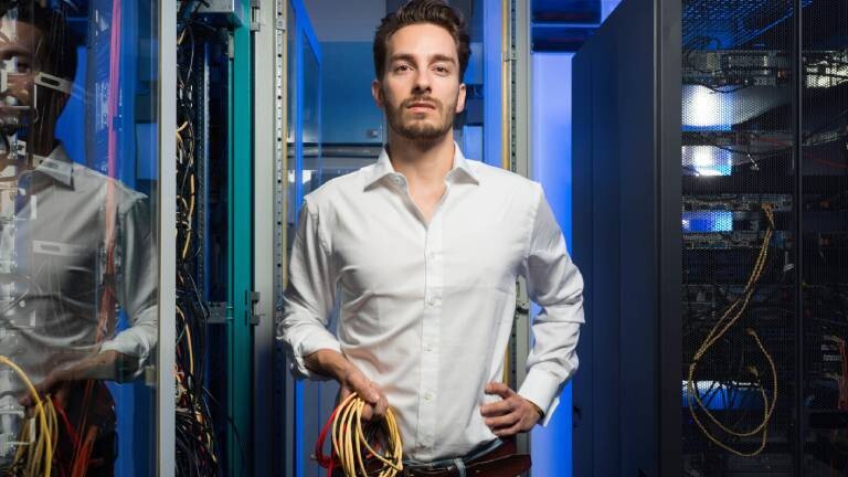 Ein Administrator einer Serverfarm steht vor der Hardware und hält bunte Kabel in der Hand.