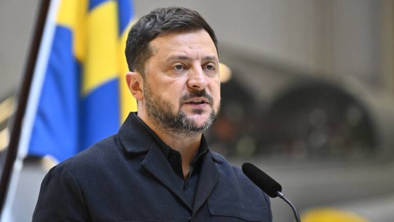 Der ukrainische Präsident Wolodymyr Selenskyj steht auf einer Pressekonferenz vor der Flagge seines Landes und spricht in ein Mikrofon.