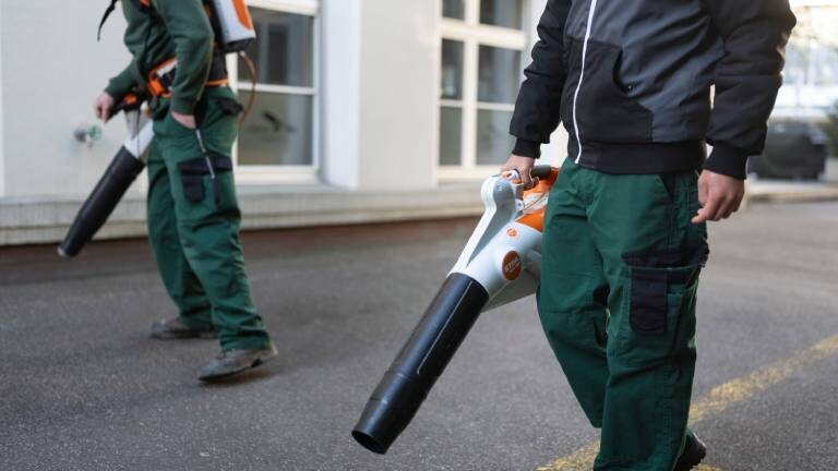 Mitarbeiter der Gartenabteilung der St. Jakob-Stiftung säubern mit elektrischen Laubbläsern den Platz um einen Bürokomplex.