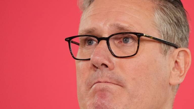 Premierminister Sir Keir Starmer hält eine Rede im Coin Street Neighbourhood Centre in Waterloo, London. Er kneift die Lippen aufeinander. Der Hintergrundfarbe ist rosa.