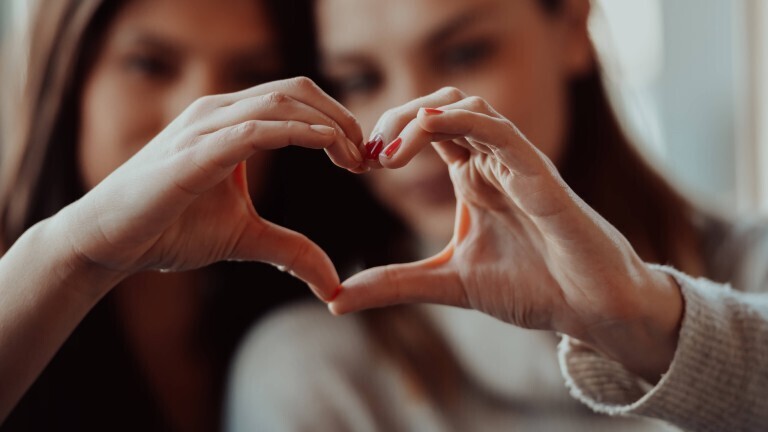 Zwei Frauen halten ihre Hände so zusammen, dass sie gemeinsam ein Herz ergeben. Die Herz-Hand ist im Vordergrund. Die beiden Frauen sind unscharf im Hintergrund.